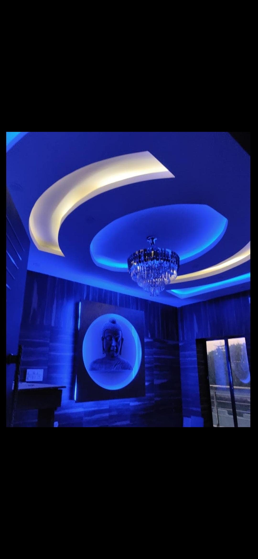 The ceiling features a modern design with curved lines illuminated by soft blue and white lighting. A crystal chandelier hangs centrally, adding a touch of elegance. A large artwork is displayed on the wall, creating a focal point in the room.