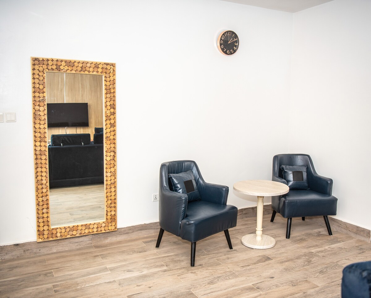 A bright living area features two stylish blue chairs positioned around a small round table. A large mirror with a decorative frame reflects the space, while a clock is mounted on the wall above. The flooring consists of light-colored wood planks.