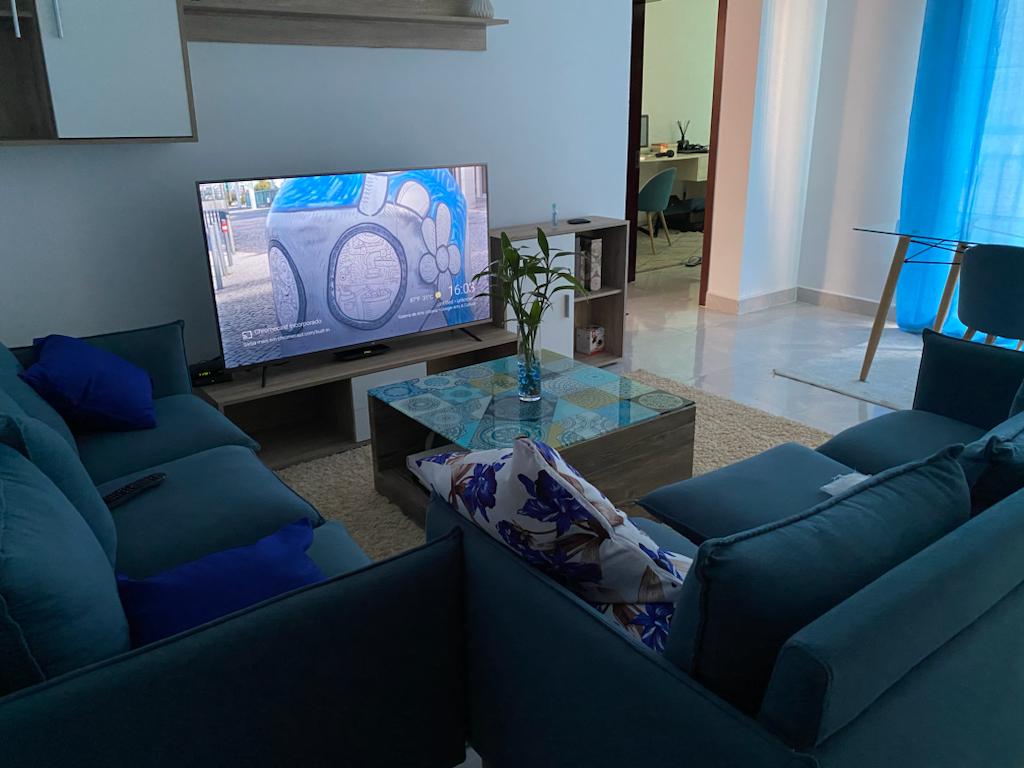 A modern living area is shown, featuring a dark blue sectional sofa and a glass coffee table adorned with decorative items. A large television displays graphics while light filters through a translucent blue curtain, illuminating the space. A nearby dining area is visible in the background.