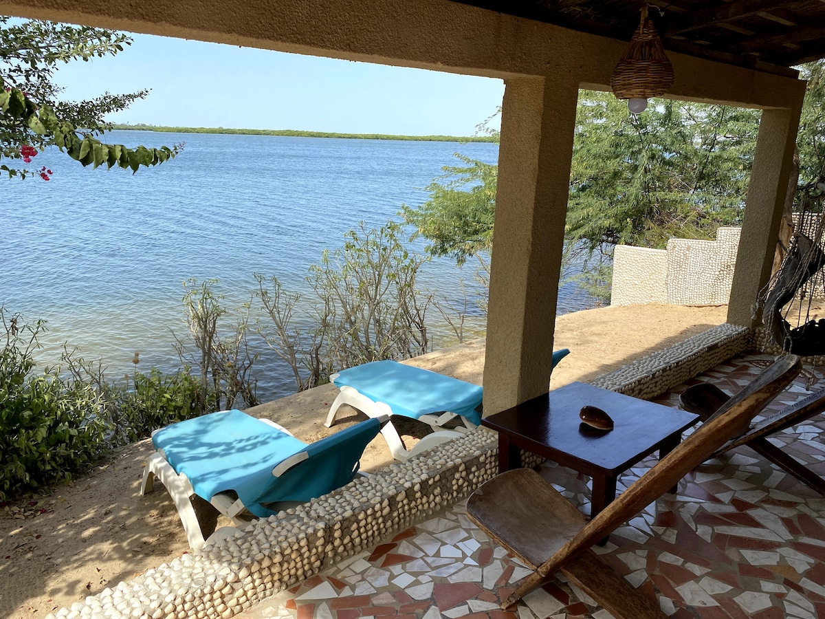 A shaded terrace overlooks the water, featuring two blue lounge chairs positioned on sandy ground. A wooden table with a decorative bowl sits nearby, while a backdrop of greenery and the tranquil water can be seen. Natural elements blend harmoniously with the outdoor space.