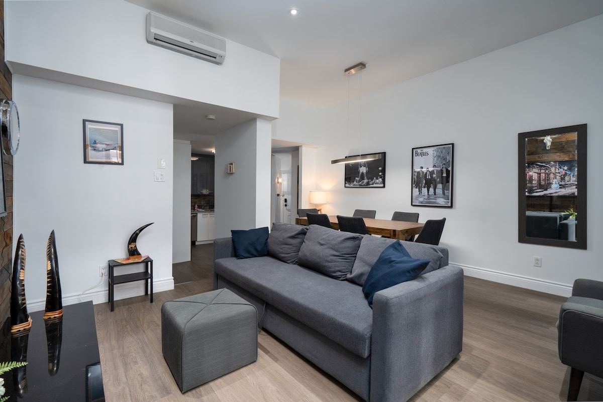 A modern living space is presented with a spacious gray sofa adorned with dark cushions. A coffee table and decorative elements add depth. A dining area with a wooden table is visible in the background, while framed artworks enhance the ambiance.