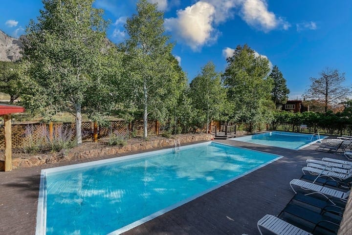 Two sparkling swimming pools surrounded by trees and landscaped greenery are visible. Sun loungers line the pool decks, inviting relaxation under a clear blue sky. A mountain backdrop enhances the serene outdoor setting.
