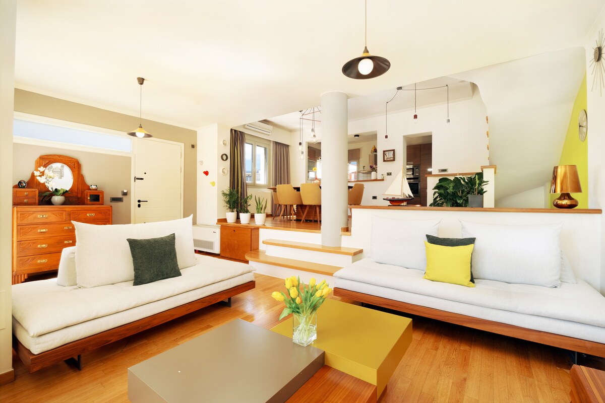 The living area is designed with two light-colored sofas and a coffee table. A vase of yellow flowers adds a touch of color. Light wood flooring complements the bright, airy atmosphere. Stairs lead to the dining area, which is visible in the background.