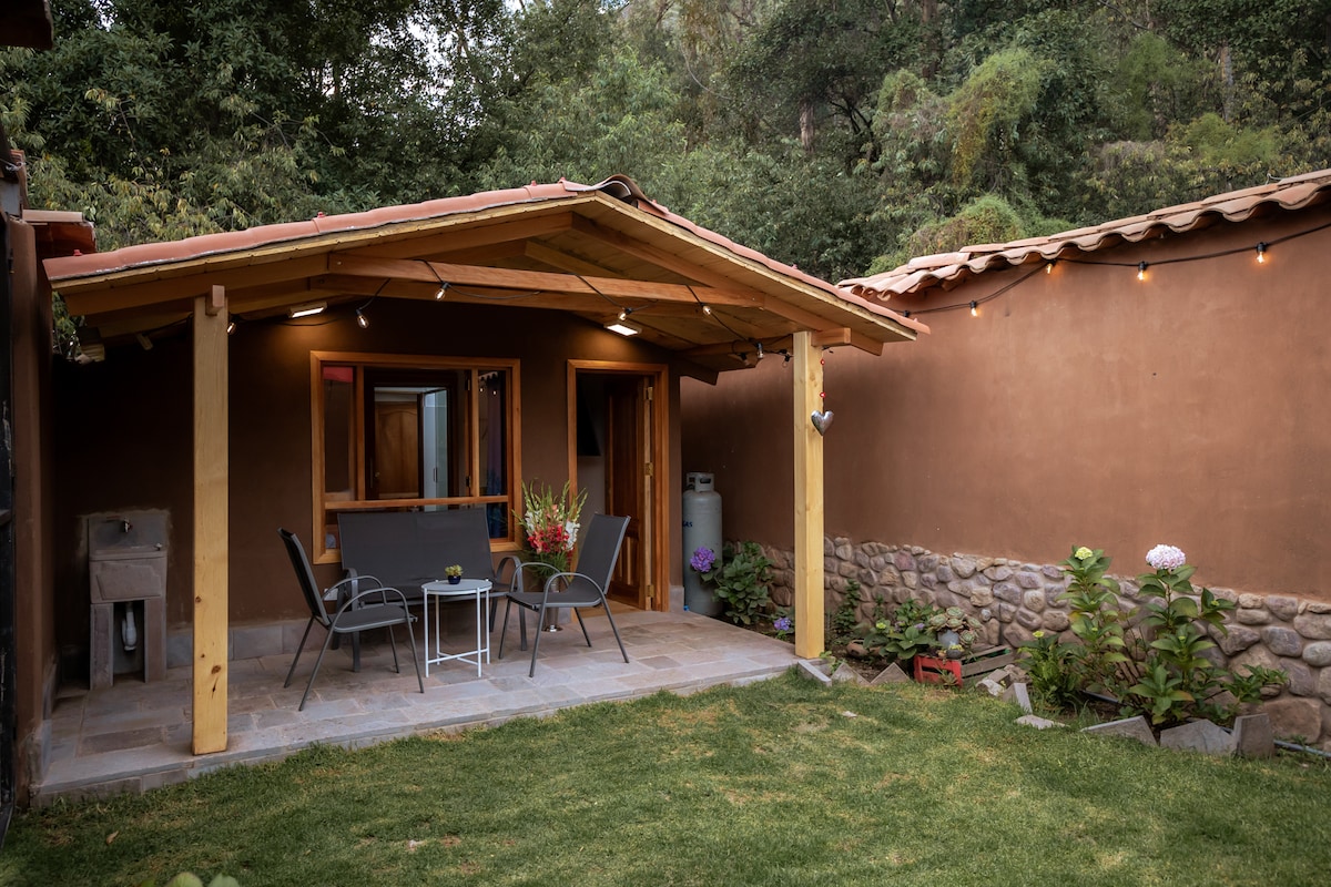 A private patio is visible, featuring a covered porch with a table and chairs. Lush greenery surrounds the area, complemented by flower beds. The entrance to the tiny house can be seen through wooden doors, set against a natural backdrop of trees.