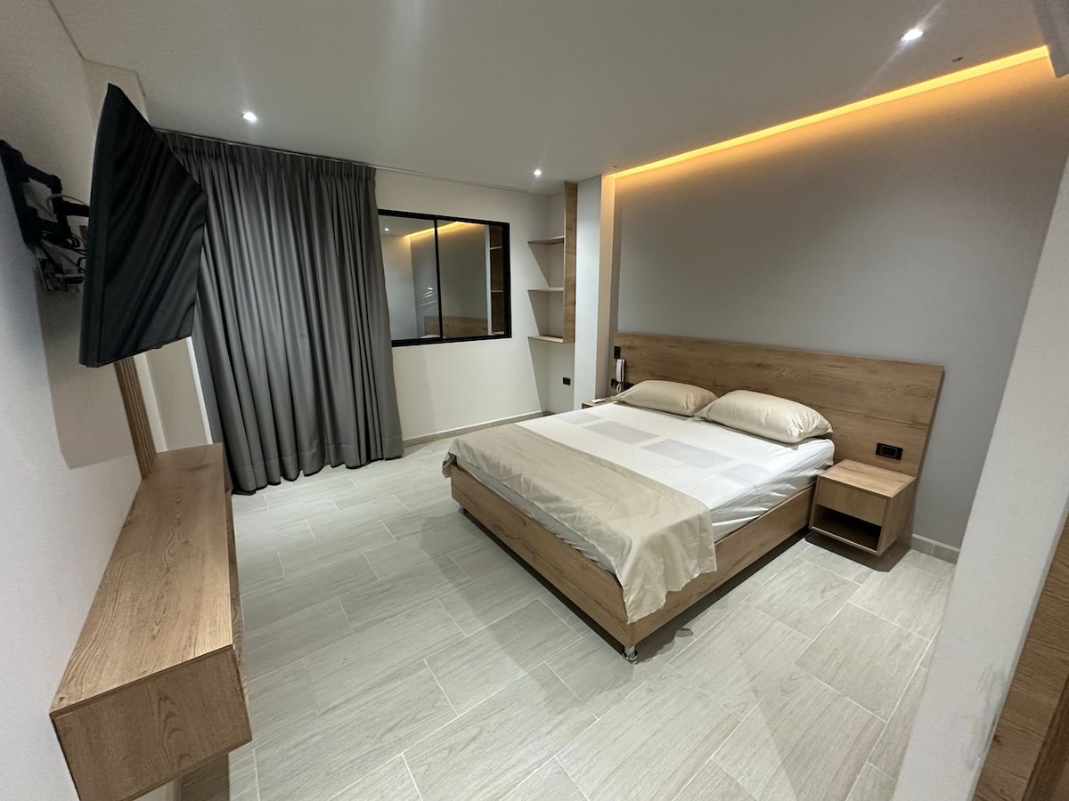 A modern bedroom features a spacious bed with neutral bedding against a backdrop of warm lighting. A wall-mounted TV is positioned opposite the bed, while a large window with draped curtains provides natural light. Wooden accents complement the sleek design.