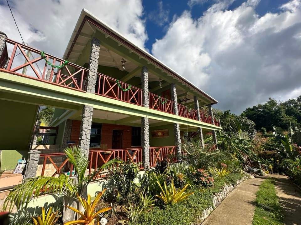 A three-story building is framed by vibrant greenery and pathways. The structure features wooden railings and stone pillars, contributing to its natural charm. A variety of tropical plants surround the exterior, enhancing its inviting presence against a backdrop of scattered clouds.