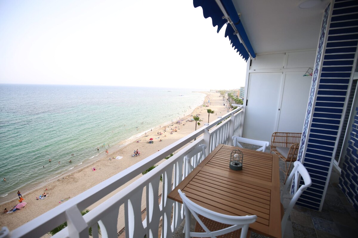 A balcony offers a view of the sandy beach and turquoise waters, with beachgoers visible below. The outdoor space features a wooden table and four white chairs, set against a backdrop of blue and white decorative railings.