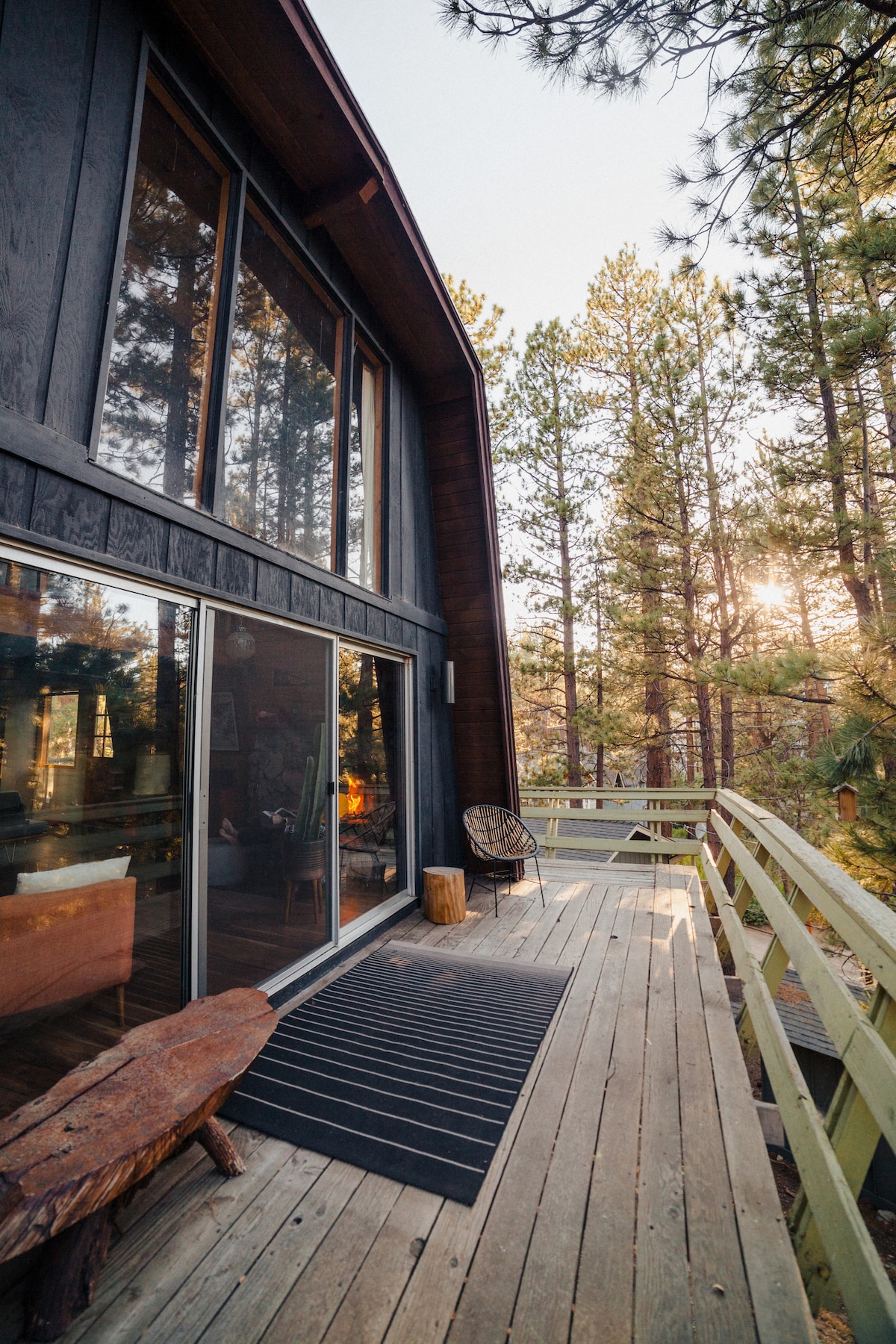 A wrap-around deck extends from the cabin, bordered by tall trees. Expansive glass doors lead into the living space, while a natural wood bench and a textured doormat provide inviting touches. Soft light filters through the forest, enhancing the cabin's outdoor charm.