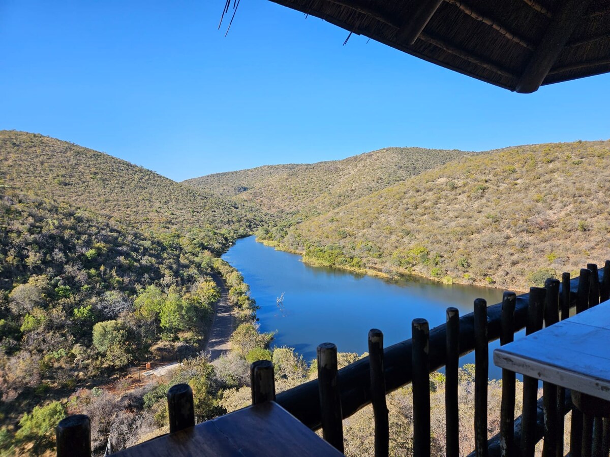 The view from a covered patio overlooks a serene river winding through lush, rolling hills. The landscape features various shades of green and brown, with gentle slopes creating a natural amphitheater. A wooden railing frames the scene, enhancing the connection to the outdoors.