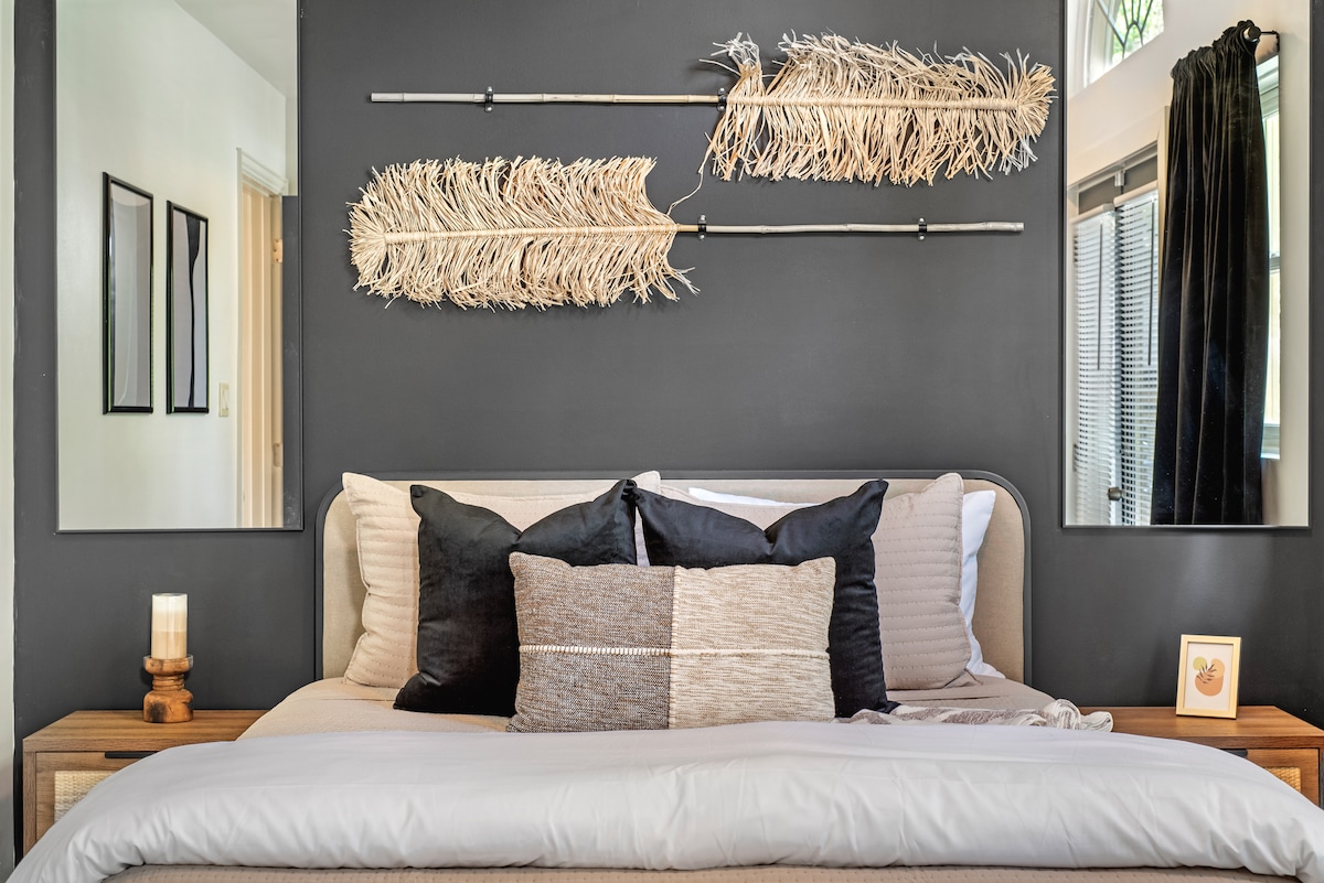 A stylish bedroom features a neutral color palette with a plush bed adorned with layered pillows. Textured wall decor made of natural materials is displayed above the headboard, while large mirrors amplify the room's light, contributing to a modern yet cozy atmosphere.