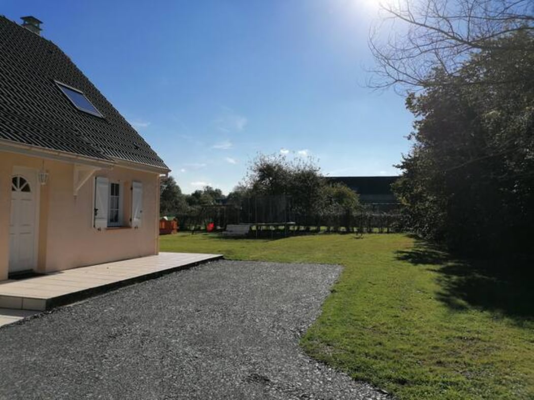 The exterior of the house is visible, highlighting a spacious garden with well-maintained grass. Sunlight bathes the area, creating a bright atmosphere. A gravel pathway leads to the entrance, and colorful play equipment can be seen in the background, indicating child-friendly outdoor space.