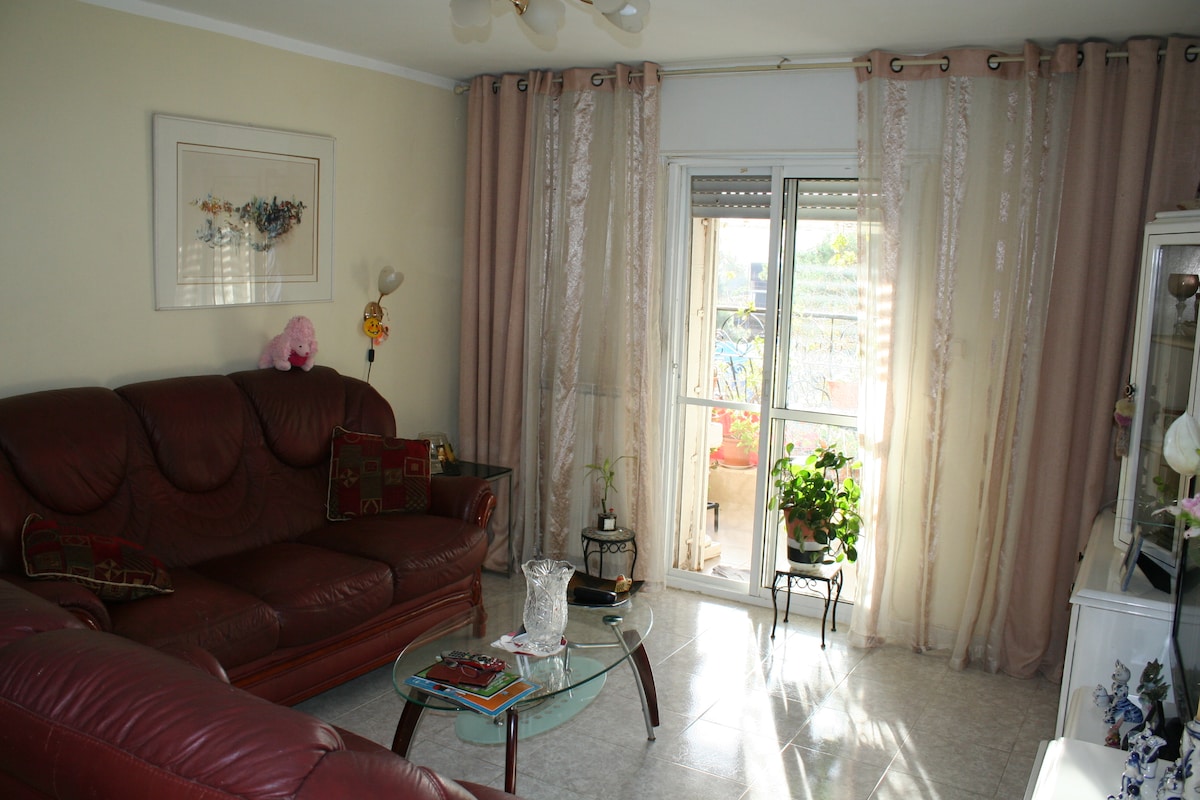 A living area features a comfortable red sofa set accompanied by a glass coffee table. Natural light enters through a large window adorned with sheer curtains, highlighting a potted plant and a small balcony area beyond. A framed artwork is displayed on the wall.