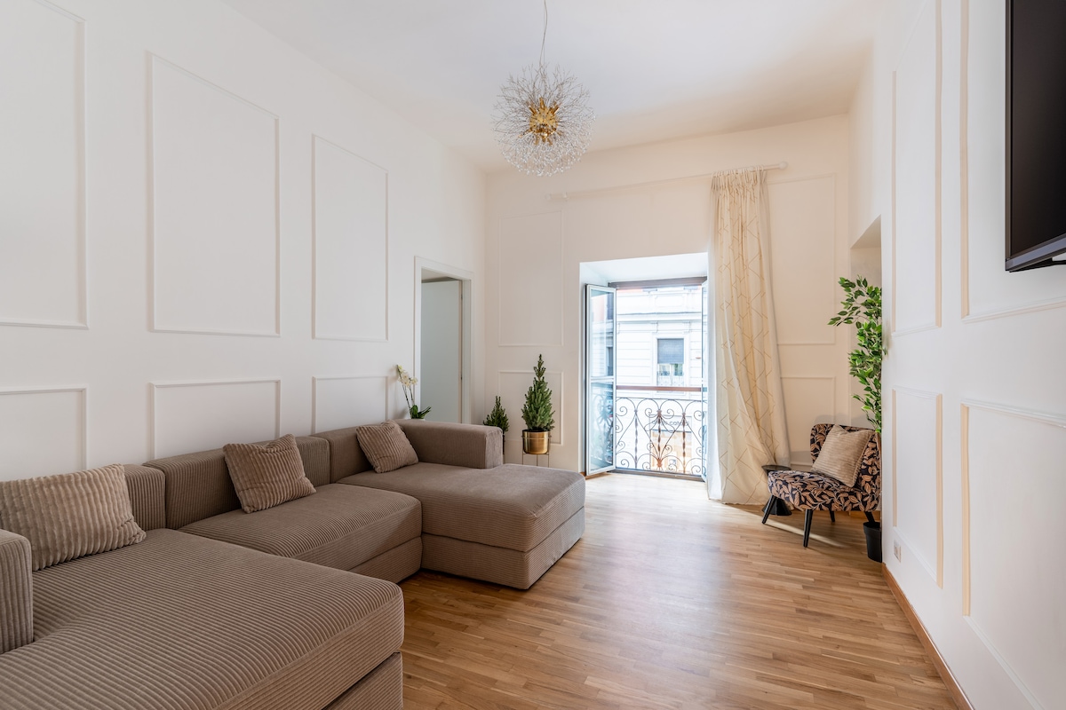 A spacious living area features a large sectional sofa, complemented by textured cushions. Natural light enters through a glass door leading to a small balcony adorned with a decorative railing. A plant adds a touch of greenery to the neutral-toned space, creating a relaxed ambiance.