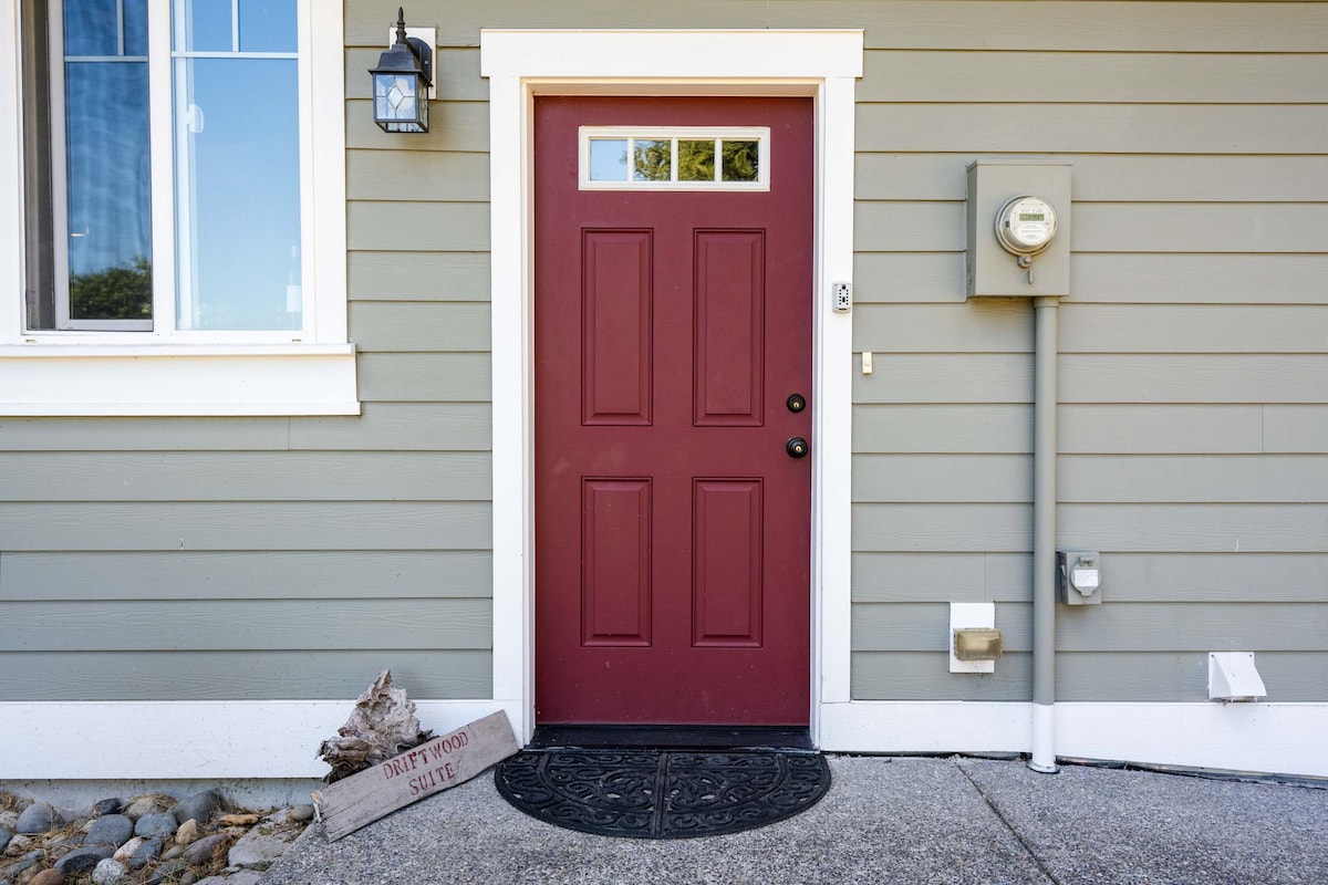 A welcoming entrance is framed by a maroon door with a decorative window above. A mat with 'Driftwood Suite' is positioned on the ground, alongside a stone pathway leading to the door. The exterior features a muted green siding and a vintage light fixture.