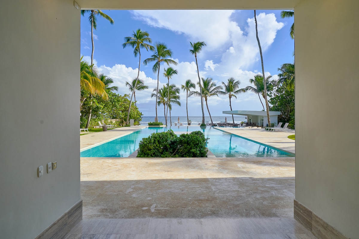 An expansive view opens from a doorway, showcasing a serene pool surrounded by palm trees and overlooking the ocean. The sky is partly cloudy, contributing to the tranquil atmosphere. The space invites relaxation in a natural setting.