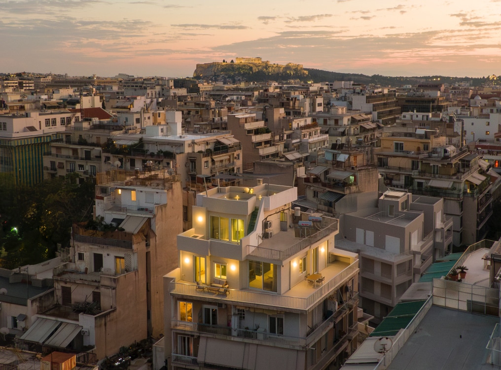 An aerial view captures the urban landscape of Athens at dusk. The foreground features a modern building illuminated from within, contrasting with the twilight sky. The Acropolis can be seen in the background, showcasing the historical significance of the city amidst a vibrant neighborhood.