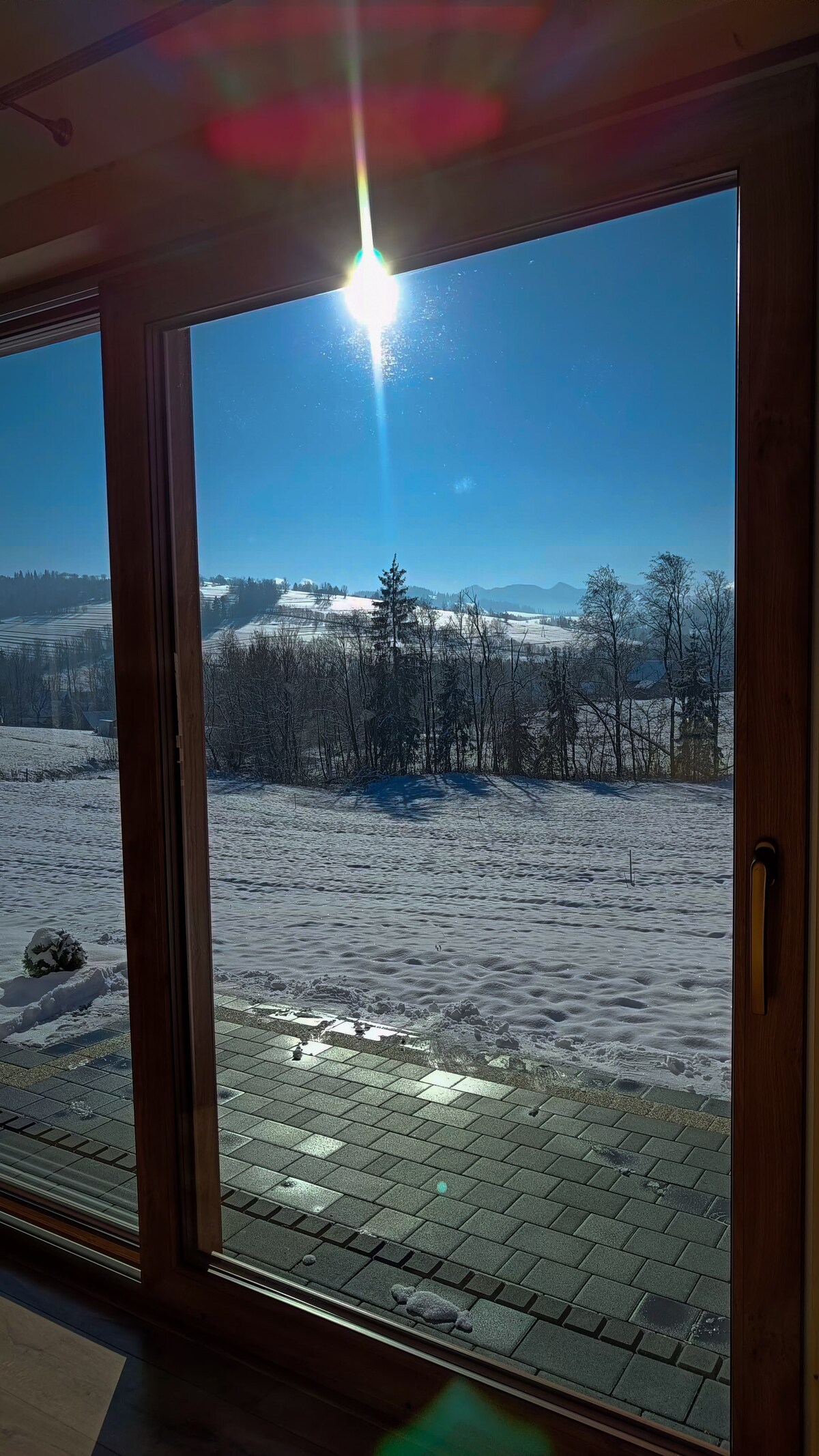 A clear view through large sliding glass doors reveals a snowy landscape, complemented by distant mountains under a bright blue sky. Sunlight glimmers on the snow-covered ground, creating a serene and inviting atmosphere.
