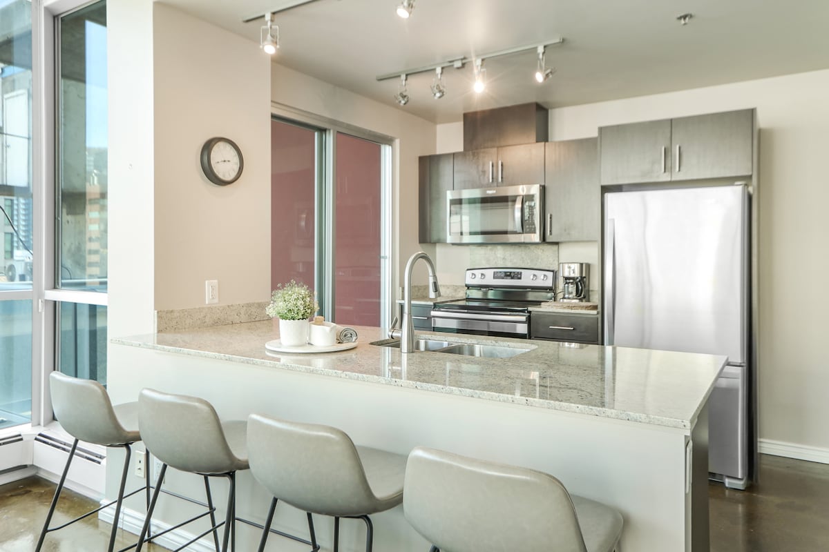 A modern kitchen is featured, equipped with stainless steel appliances including a microwave and refrigerator. A spacious granite countertop serves as a dining area with four stools arranged neatly. Natural light streams in through large windows, enhancing the open atmosphere.