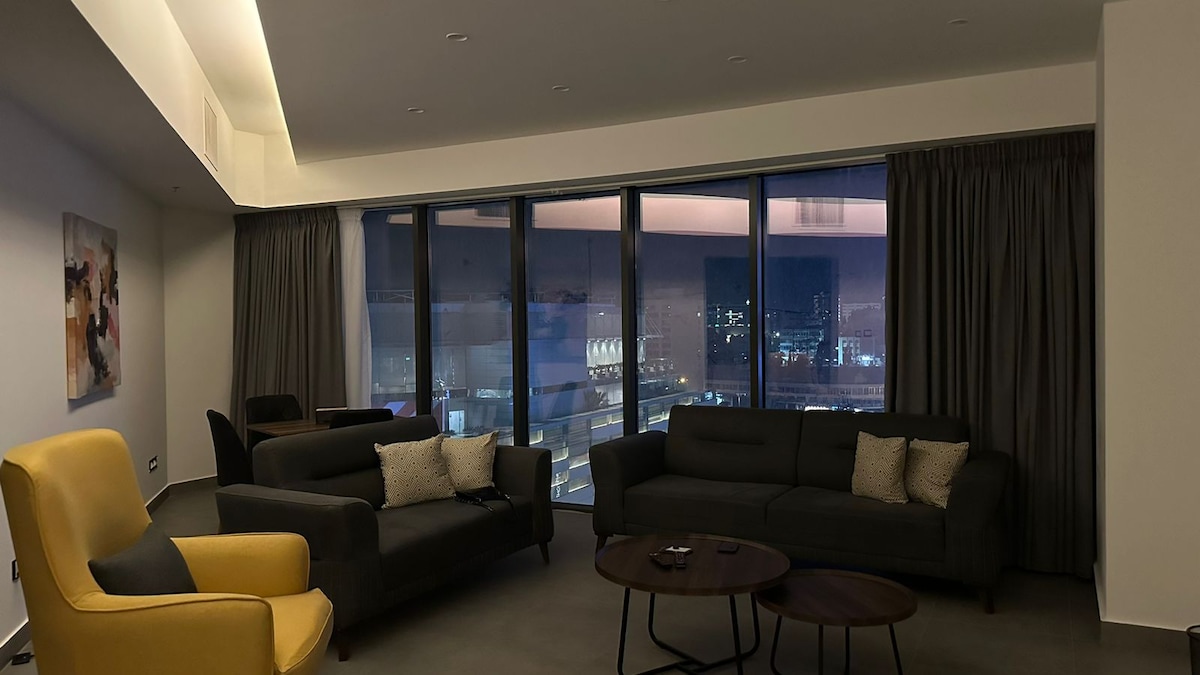 The living area features large floor-to-ceiling windows that offer a view of the city skyline at dusk. Two dark sofas are arranged around a circular coffee table, complemented by a yellow accent chair. Soft lighting enhances the modern decor, creating a cozy ambiance.