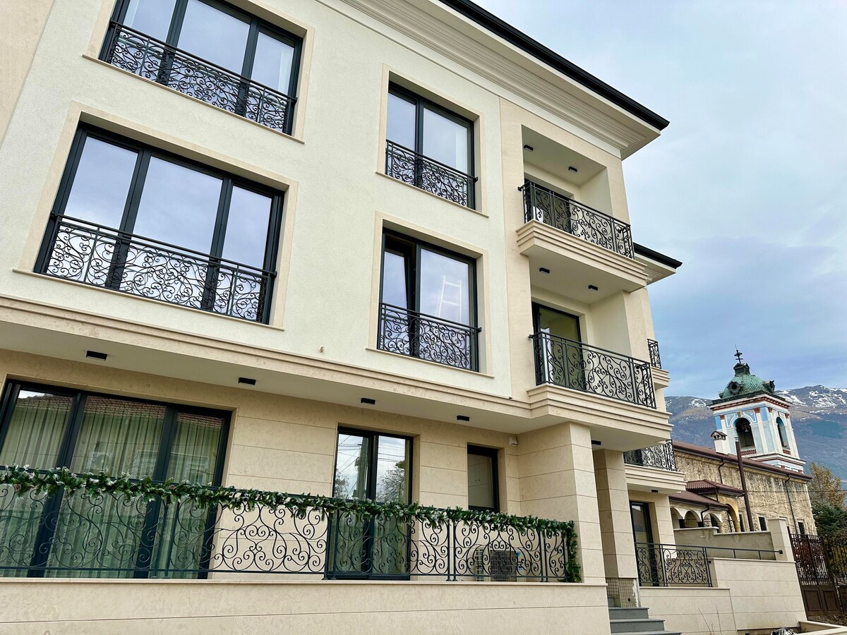 A three-story building features elegant balconies adorned with intricate wrought iron railings. Large windows provide ample natural light, while the surrounding area is framed by distant mountains. A decorative structure is visible, adding to the architectural interest of the environment.