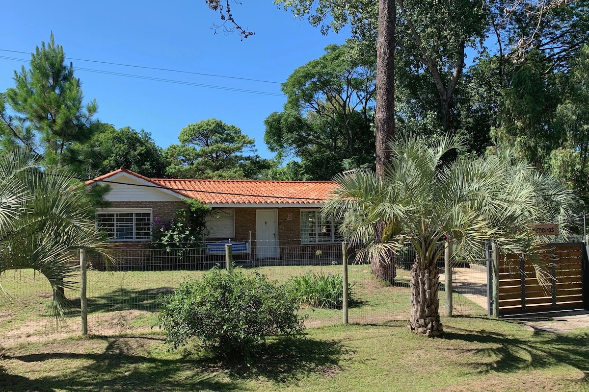 A single-story brick house is surrounded by lush greenery and palm trees. The exterior features a red-tiled roof and large windows. A fenced yard offers a sense of privacy, with a pathway leading to the entrance. The peaceful setting is enhanced by the natural environment.
