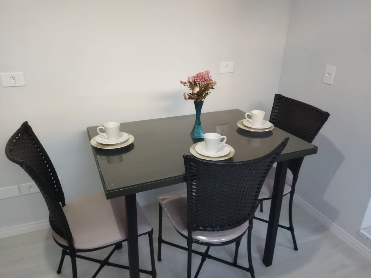 A dining table set for two features a sleek black top and is surrounded by four wicker chairs. Two cups and plates are neatly arranged on the table, accompanied by a small vase with flowers, enhancing the dining area.