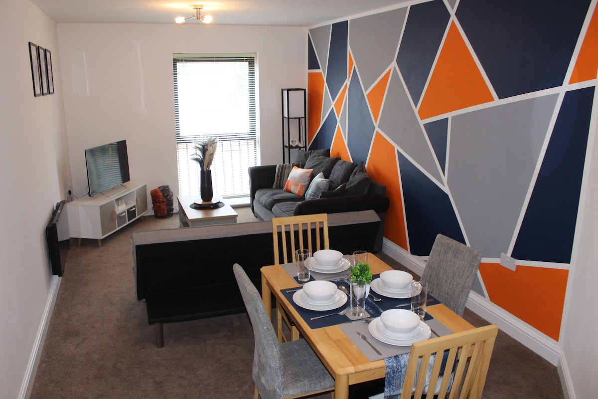 An open plan living and dining area is showcased, featuring a grey sectional sofa with decorative pillows. A wooden dining table is set for four, complete with plates and utensils. Geometric wall designs in grey, blue, and orange add a modern touch, while a balcony is visible through the window.