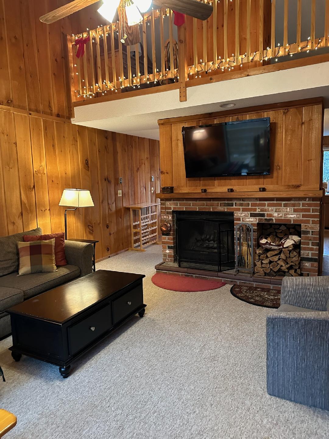 A warm living area features wooden paneled walls and cozy furnishings. A simple black coffee table sits in front of a comfortable sofa and two armchairs. A brick fireplace with stacked logs provides a focal point, with a flat-screen TV mounted above.