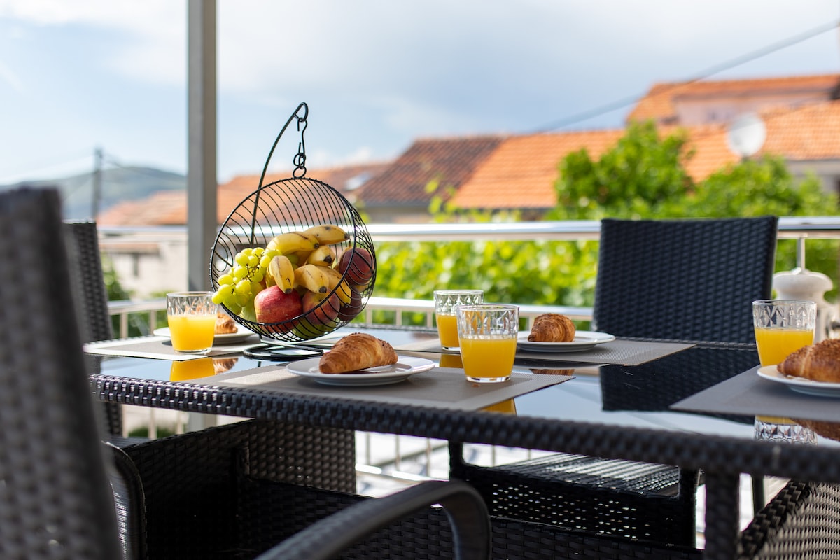 A dining table is set on a large terrace, featuring wicker chairs and a dark table. A basket of fresh fruits is displayed prominently. Glasses of orange juice and croissants are arranged neatly, enhancing the inviting setting for a meal outdoors. Lush greenery is visible in the background.