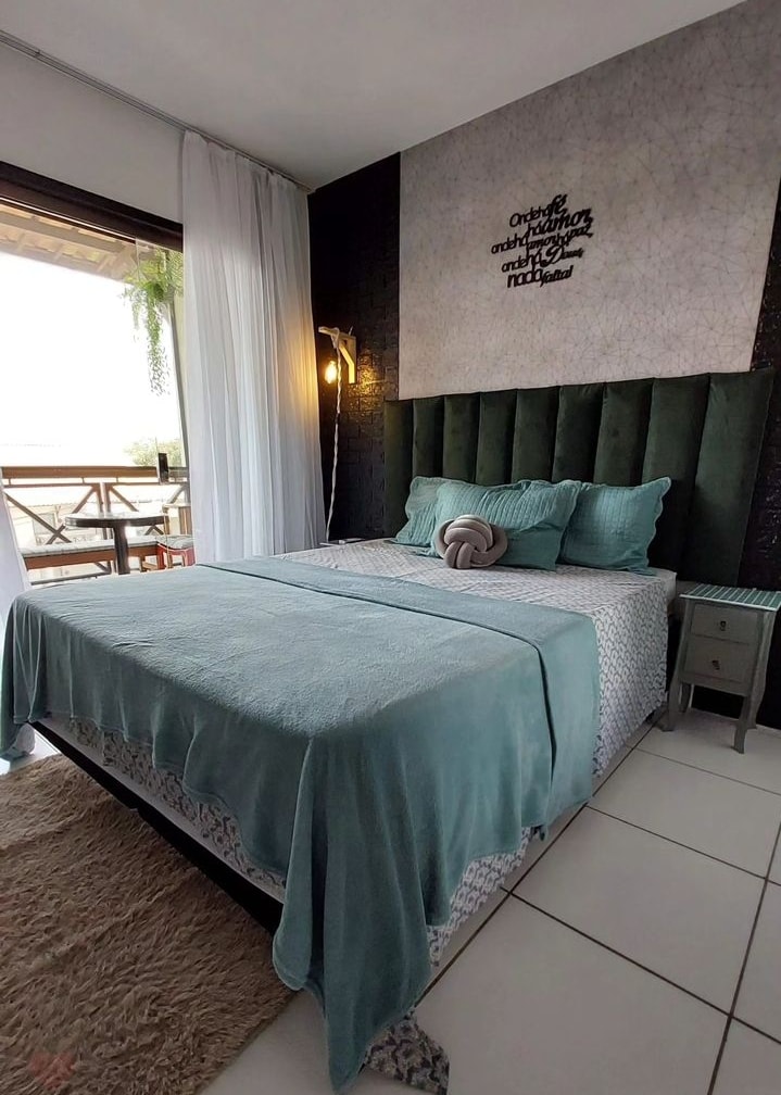 A stylish bedroom features a spacious bed with a soft teal blanket, accented by coordinating pillows. A textured wall behind the bed adds interest, while large windows allow natural light to fill the room. A small bedside table and decorative rug complete the inviting space.