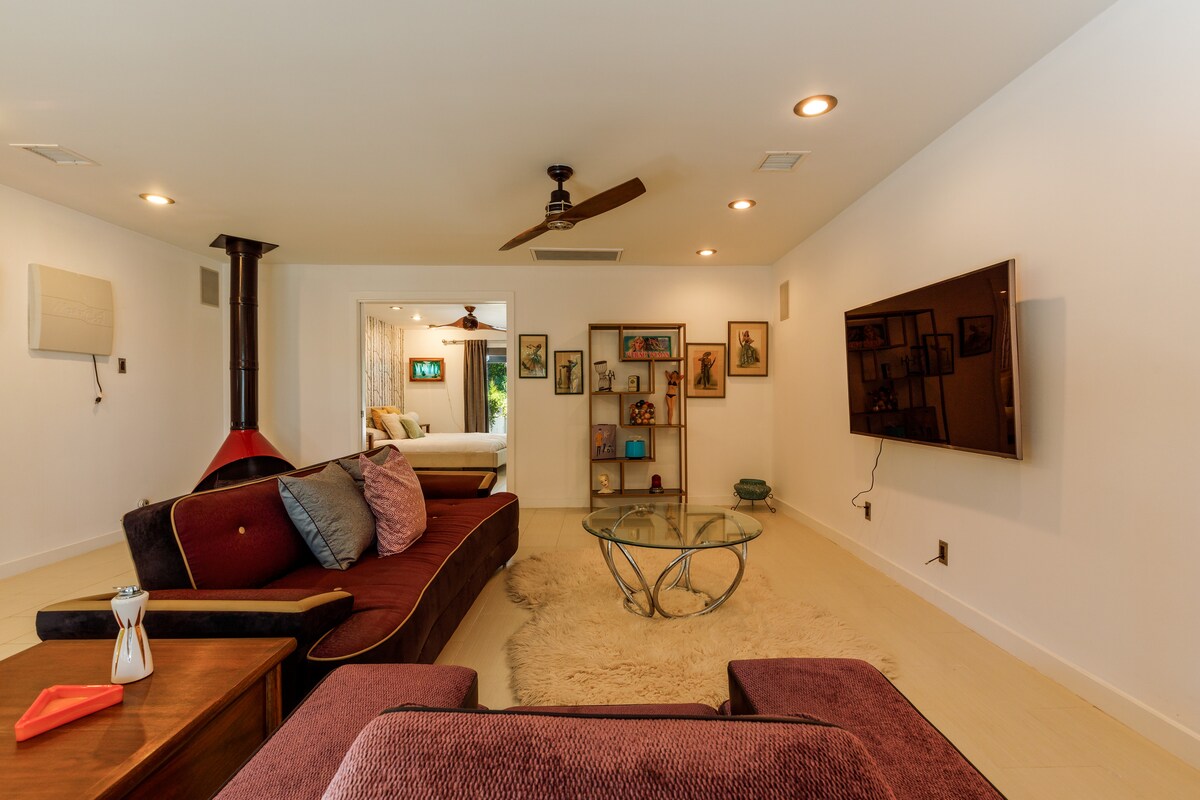 A spacious living area features a contemporary glass coffee table and a plush sofa with vibrant throw pillows. An entertainment unit with a large flat-screen television is mounted on the wall. A cozy electric fireplace adds warmth to the room, enhancing its welcoming ambiance.