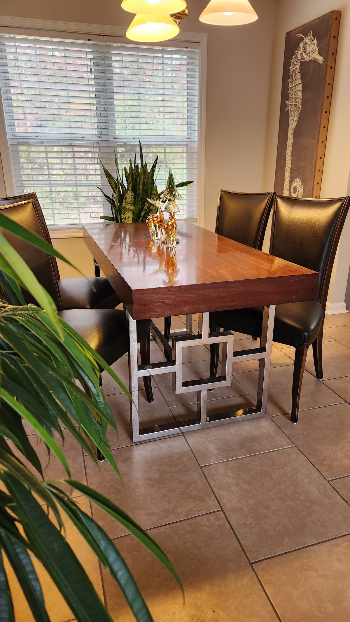 A dining area features a sleek rectangular table with a wooden top and a geometric metal base. Four black leather chairs are arranged around the table. Lush green plants are positioned on the table and nearby, providing a touch of natural decor. Soft light filters through the window.