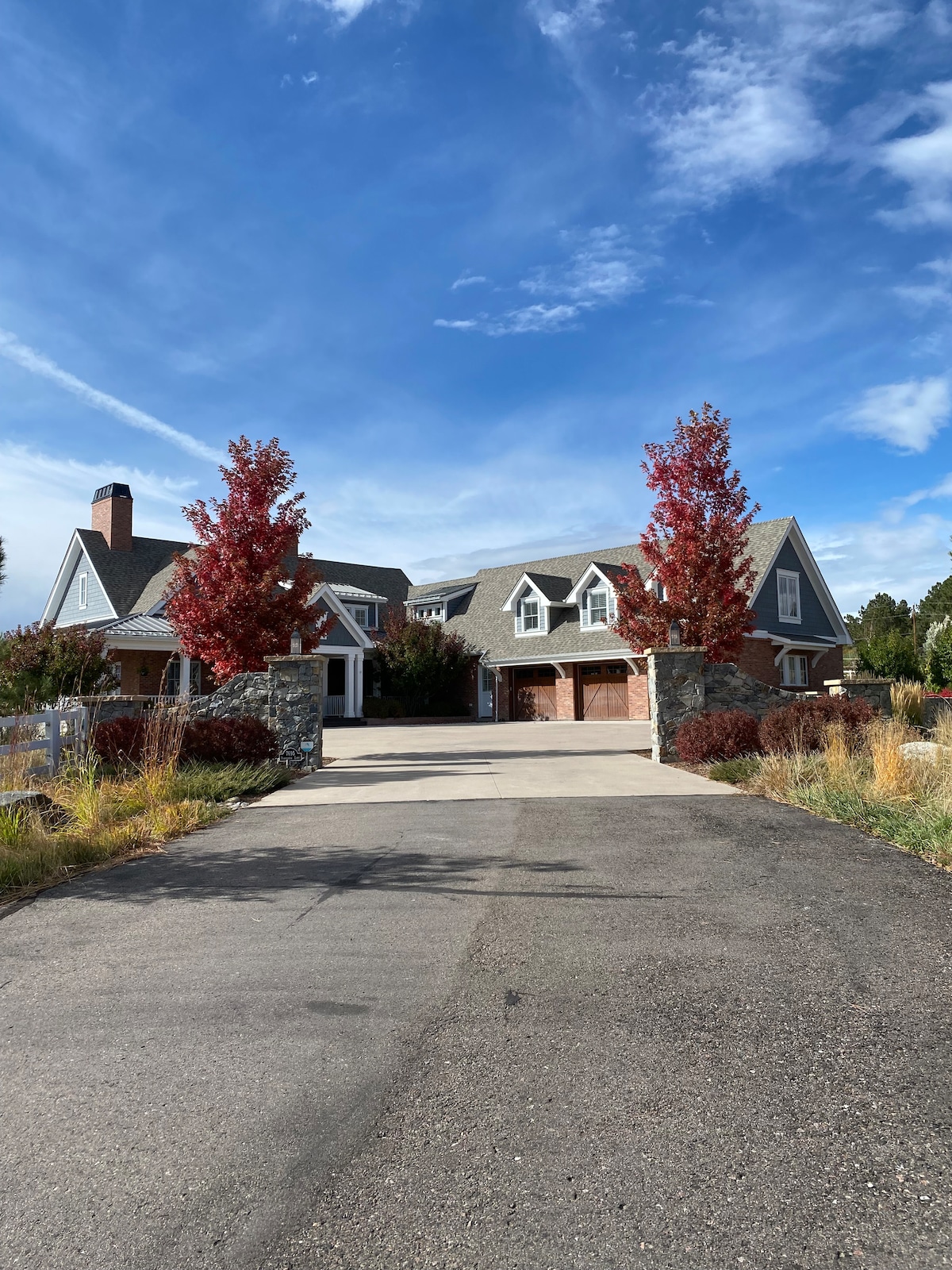 The image shows a grand home with a sloped roof, flanked by vibrant red trees. A well-maintained driveway leads to the entrance, highlighting the stone pillars at the sides. A spacious yard surrounds the house, enhancing the inviting exterior.