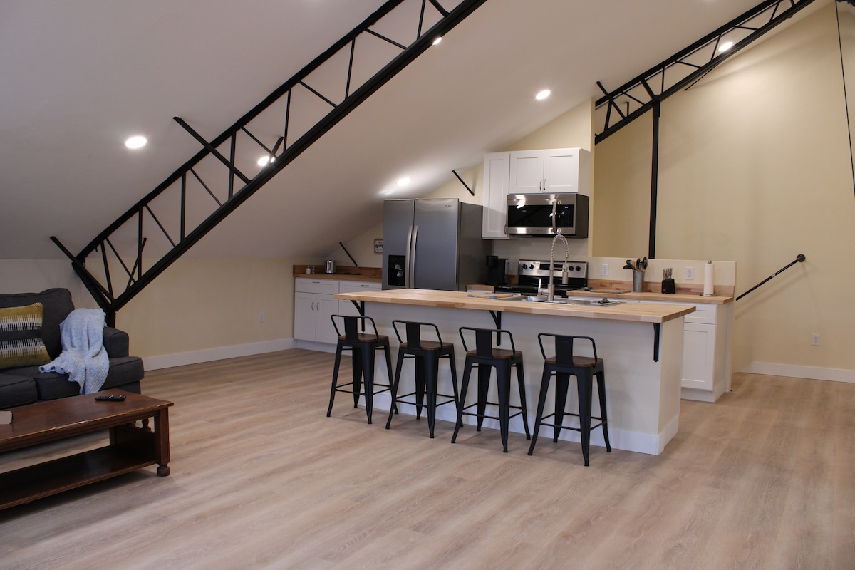 An open concept kitchen and living area is presented with a spacious island featuring seating for six. Stainless steel appliances are visible alongside modern cabinetry. Natural light is provided by overhead lighting and large windows, enhancing the airy feel of the space.