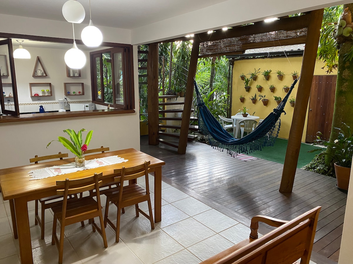 An open dining area features a wooden table with four chairs, complemented by a bench. Behind, a cozy outdoor space is highlighted by greenery, a hammock, and a small seating area. Large windows provide a view into the kitchen, enhancing the airy feel.