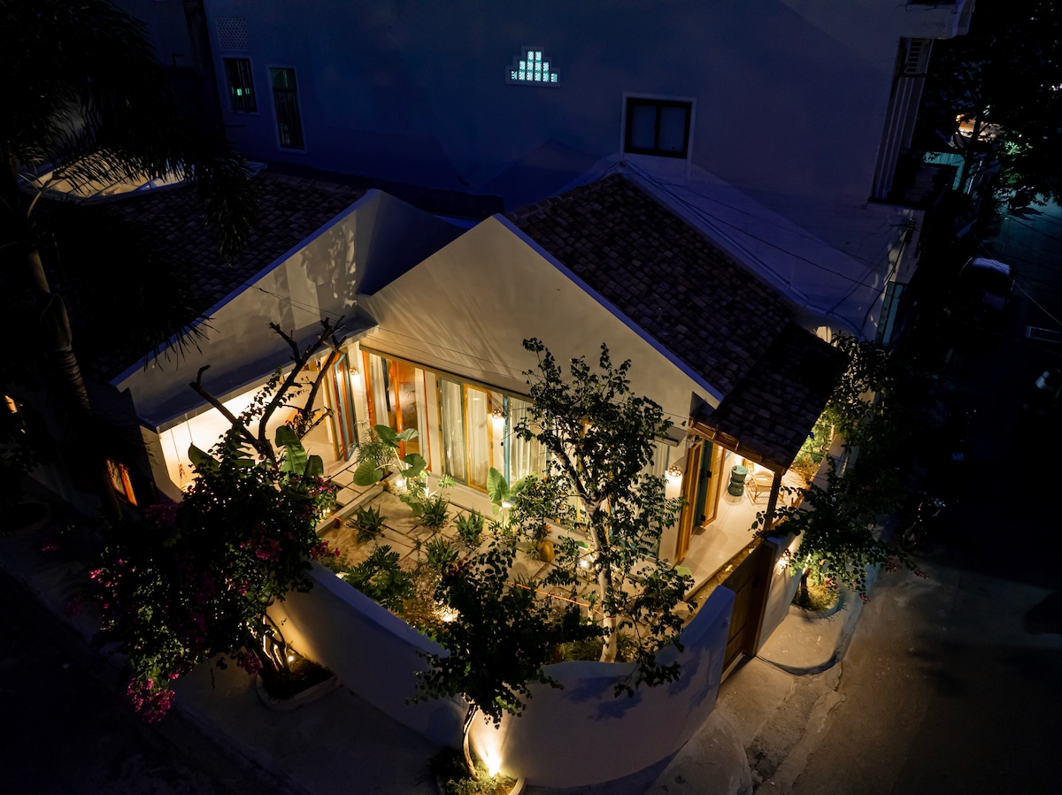 An aerial view of the villa showcases its unique architecture, blending with the outdoor garden. Warm lighting highlights the greenery and wooden features, while the inviting exterior creates a welcoming atmosphere. The surrounding area reflects local charm, enhancing the overall setting.