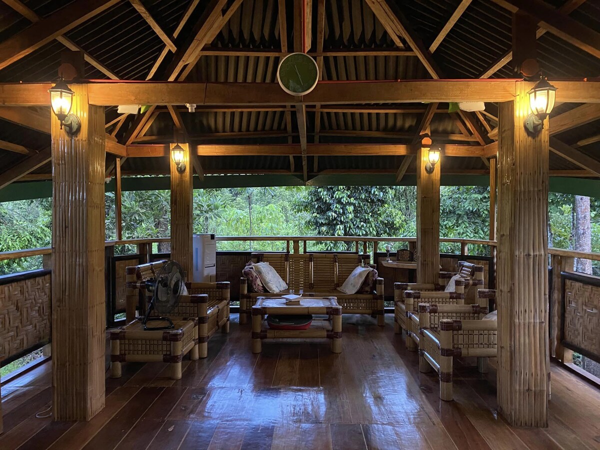An open pavilion features a wooden structure with bamboo furnishings, surrounded by lush greenery. Several seating arrangements are available, including a central table. Illuminated by warm light fixtures, the space invites relaxation in a natural setting.