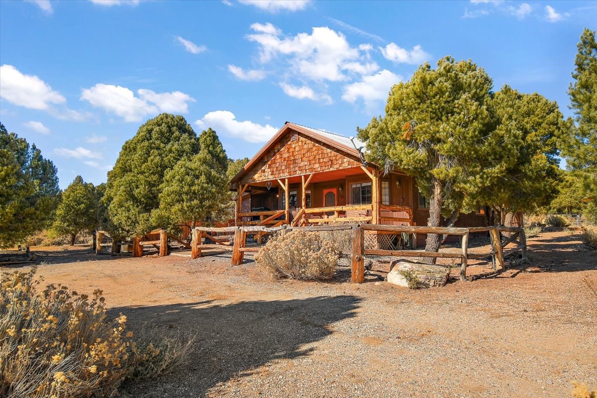 A ranch home is nestled among trees, surrounded by a gravel area. A wooden fence outlines the property, and a welcoming porch extends across the front, showcasing natural wood details. The landscape features shrubs and natural vegetation under a sunny sky with scattered clouds.