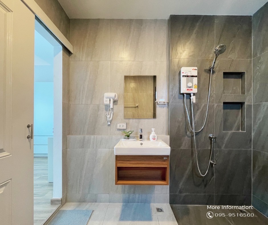 A modern bathroom features textured gray tiles and a wooden countertop under a mirror. A wall-mounted showerhead and handheld shower are arranged within the tiled shower area. Soap dispensers and a hand dryer are visible, ensuring functionality and convenience.