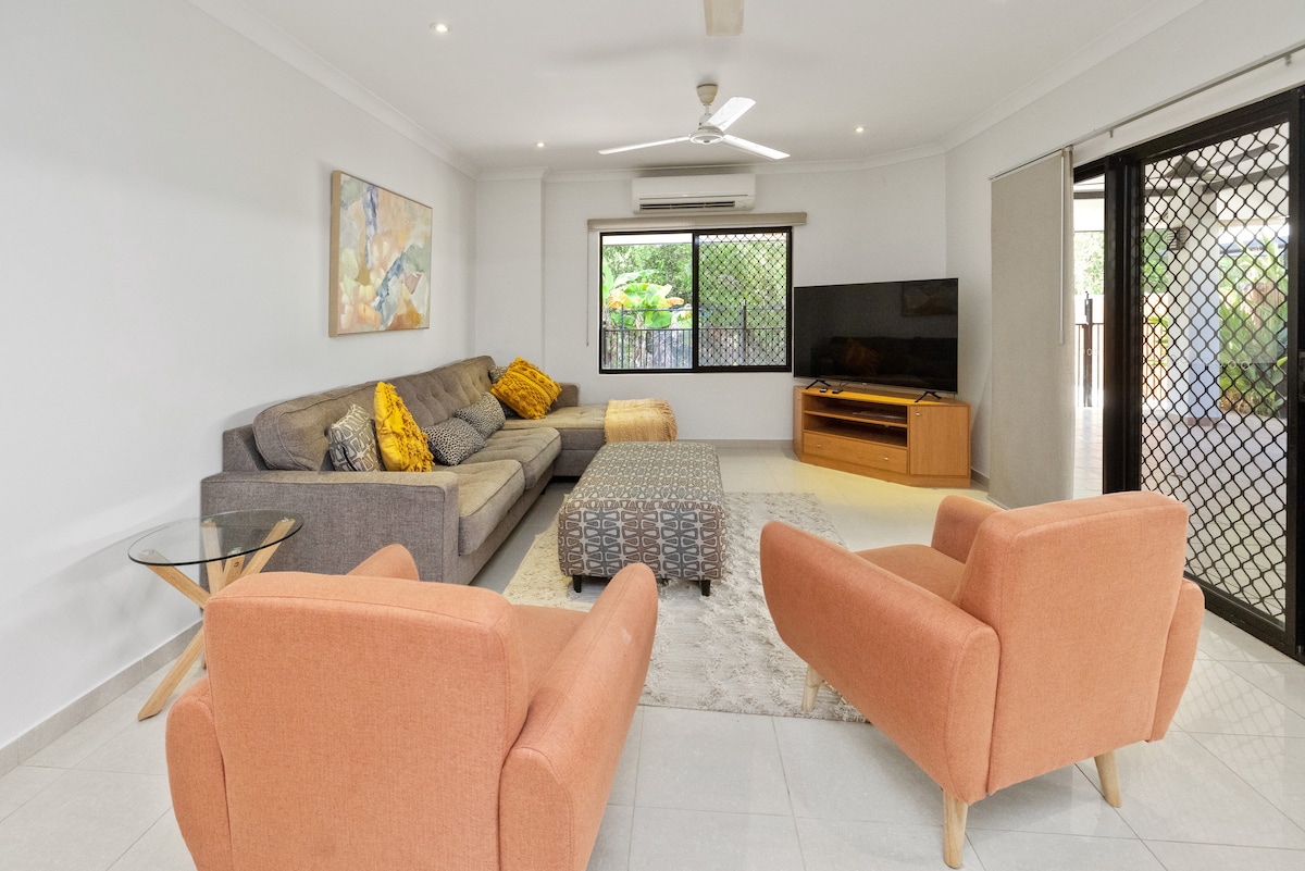 A spacious living area features a large, comfortable sofa adorned with bright cushions. Two coral armchairs are positioned nearby, and a rectangular coffee table sits in the center. Natural light filters in through large windows, adding warmth to the neutral palette.
