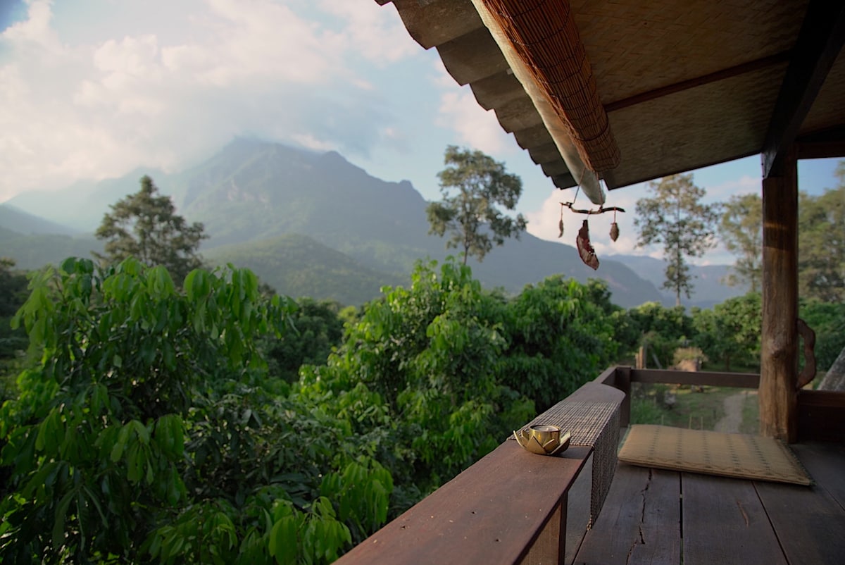 A balcony view presents lush green trees and distant mountains under a cloudy sky. A decorative wind chime hangs from the roof edge, while a small dish rests on the wooden railing. The tranquil setting invites the sounds and sights of nature.