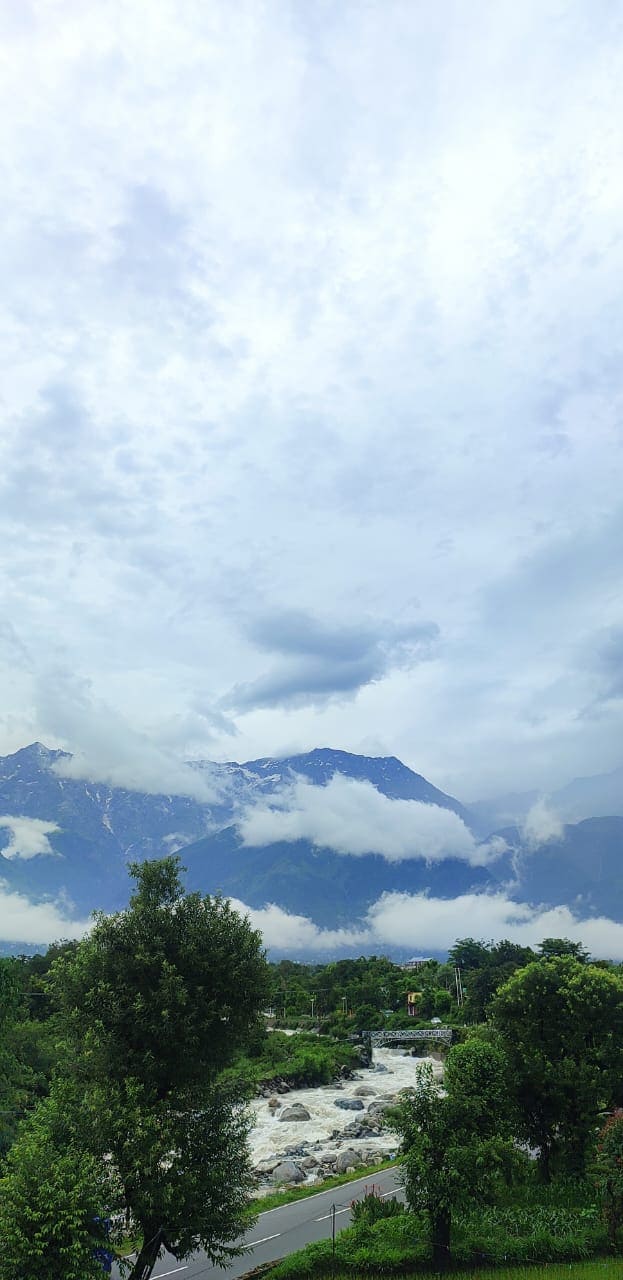 A scenic view features rolling mountains partially shrouded in clouds, while a flowing river is visible in the foreground. Lush greenery, including trees and shrubs, frames the landscape, creating a sense of tranquility and connection to nature.