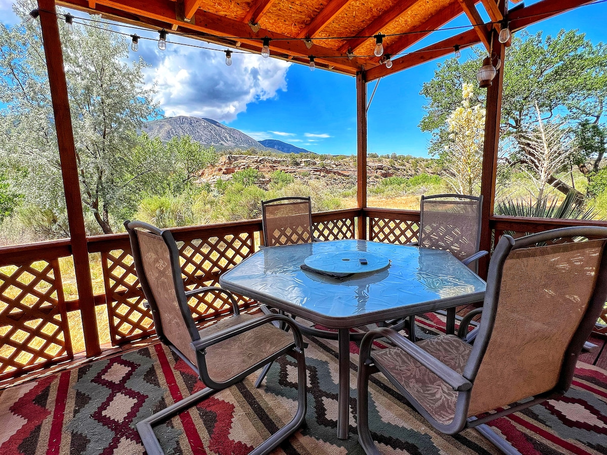 A shaded outdoor patio features a hexagonal table surrounded by six chairs, ideal for enjoying meals or relaxation. String lights hang above, enhancing the space. The view includes expansive greenery and the backdrop of rugged mountains under a clear blue sky.