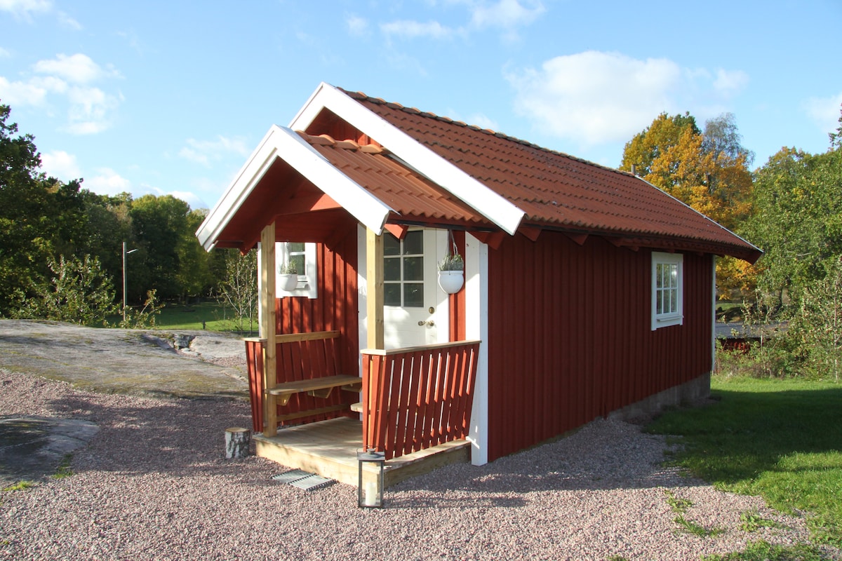 A charming tiny cottage is showcased with a red wooden exterior and a tiled roof. A small porch with steps is visible, inviting guests to the entrance. Surrounding greenery and trees complement the serene setting, while a gravel path leads up to the cottage.