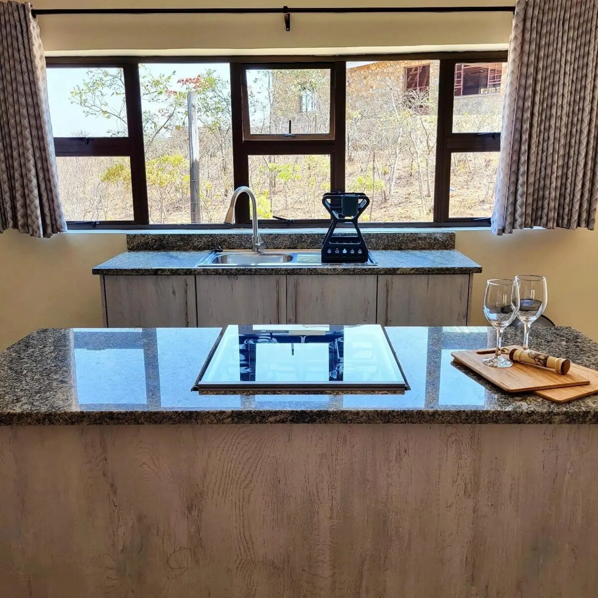 A modern kitchen is showcased with a spacious island featuring a stovetop and granite countertops. A sleek sink and high-backed faucet are positioned near a large window, allowing natural light in. Glasses and a serving board, along with a corkscrew, are arranged on the countertop.