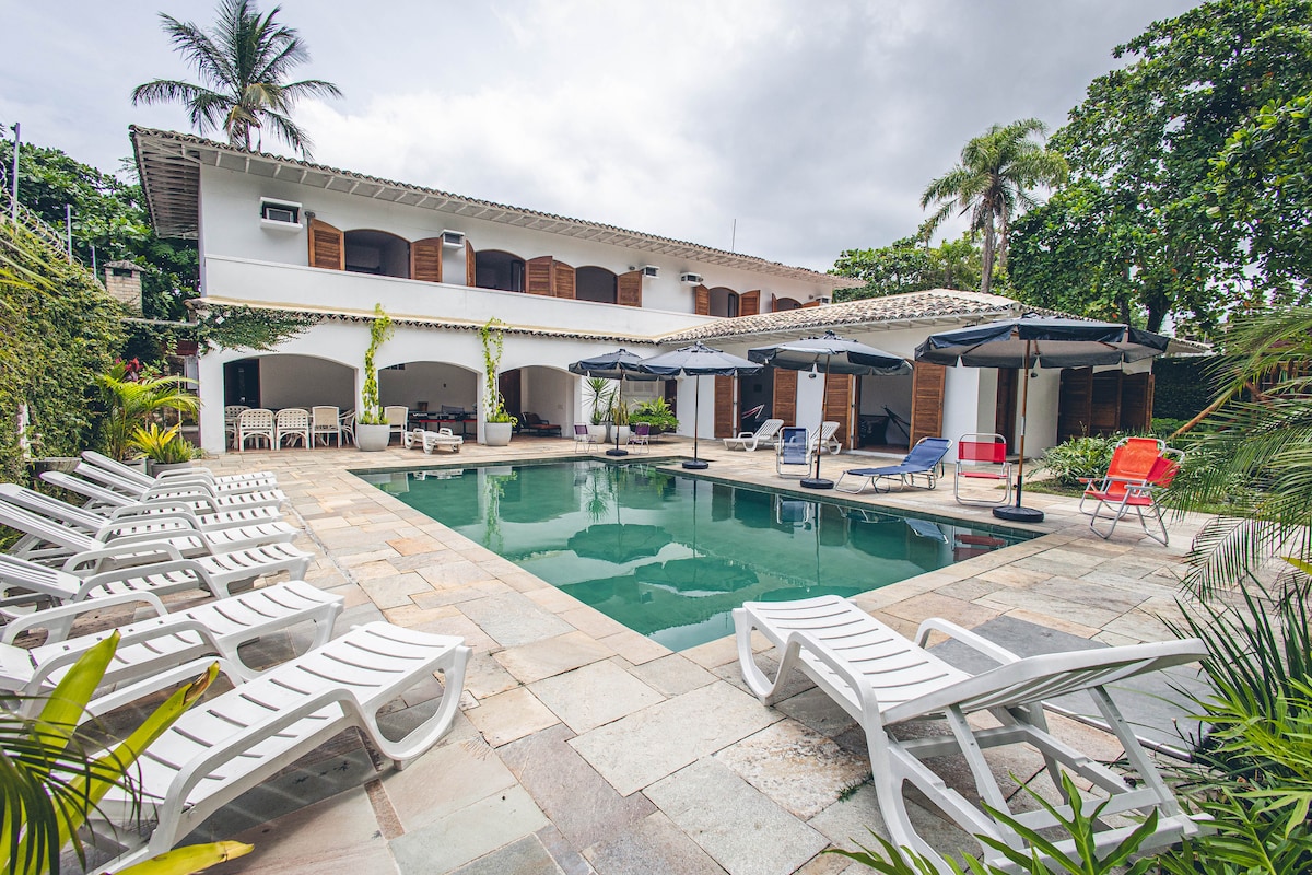 A spacious outdoor area features a swimming pool surrounded by lounge chairs, complemented by umbrellas for shade. The two-story house is partially visible, showcasing wooden shutters and lush greenery encircling the space, enhancing the feeling of privacy and tranquility.
