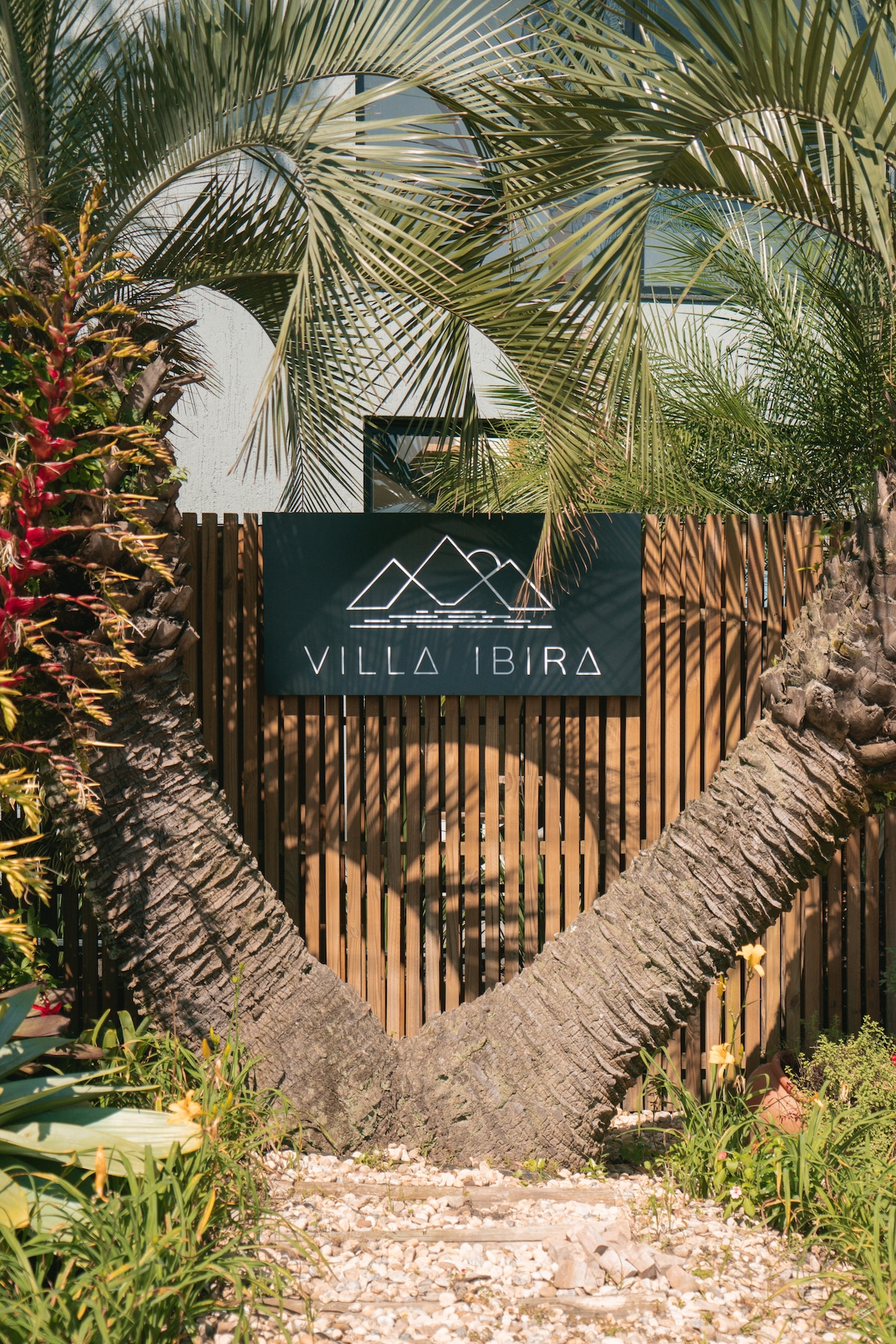 A welcoming entrance to Villa Ibira is framed by two palms, creating a natural archway. The modern sign displaying the property name is prominently featured against a backdrop of lush greenery and natural textures, inviting guests into a serene atmosphere.