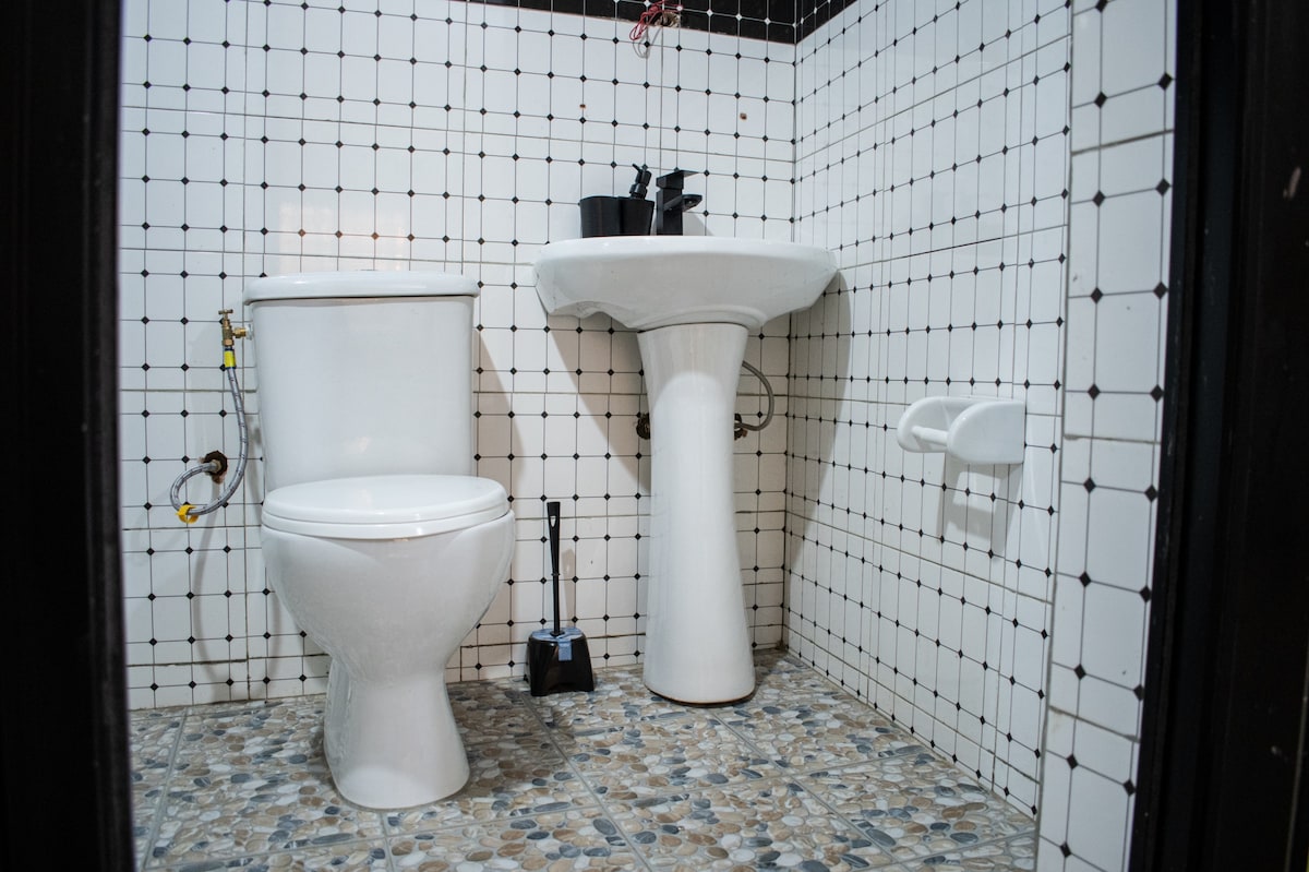 A clean bathroom displays a white toilet and a pedestal sink, both set against a backdrop of white tiles accented with black grout. The floor is composed of small, pebbled stones, providing a textured contrast to the tiled walls.
