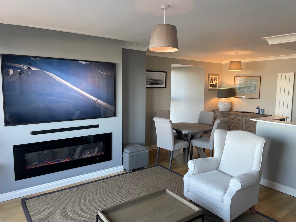A living area is presented with a large flat-screen television mounted on the wall, complemented by an electric fireplace below. A light-colored armchair and a coffee table sit atop a textured rug, while a dining table with chairs occupies a separate space, illuminated by natural light.