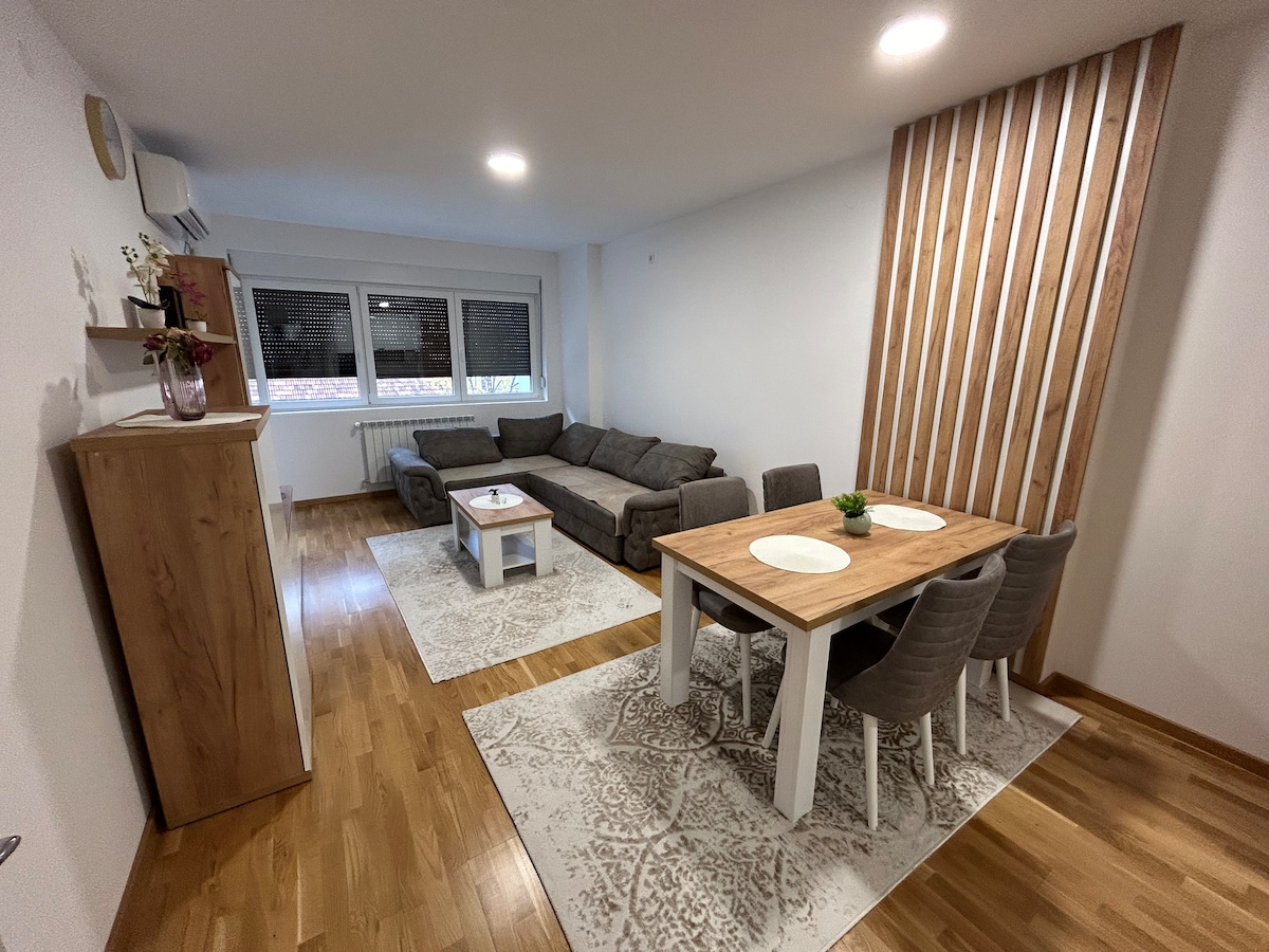 A cozy living space features a spacious gray sofa and a designated dining area with a wooden table set for four. The room is illuminated by recessed lighting and has light hardwood flooring, complemented by decorative rugs. Large windows provide natural light.